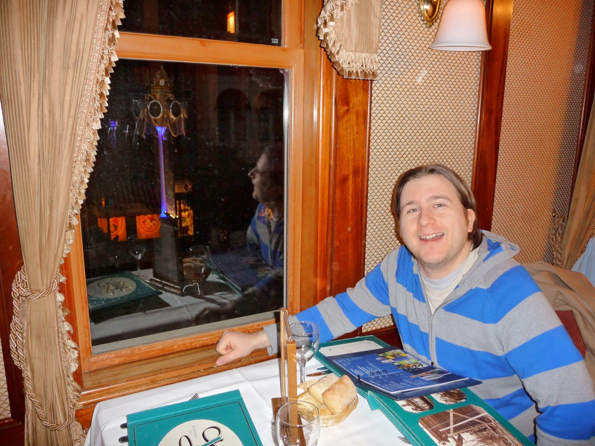 image shows a young man seated in the upstairs window of walt's restaurant in disneyland paris. He is reading the teal coloured menu and has bread rolls on the table. 