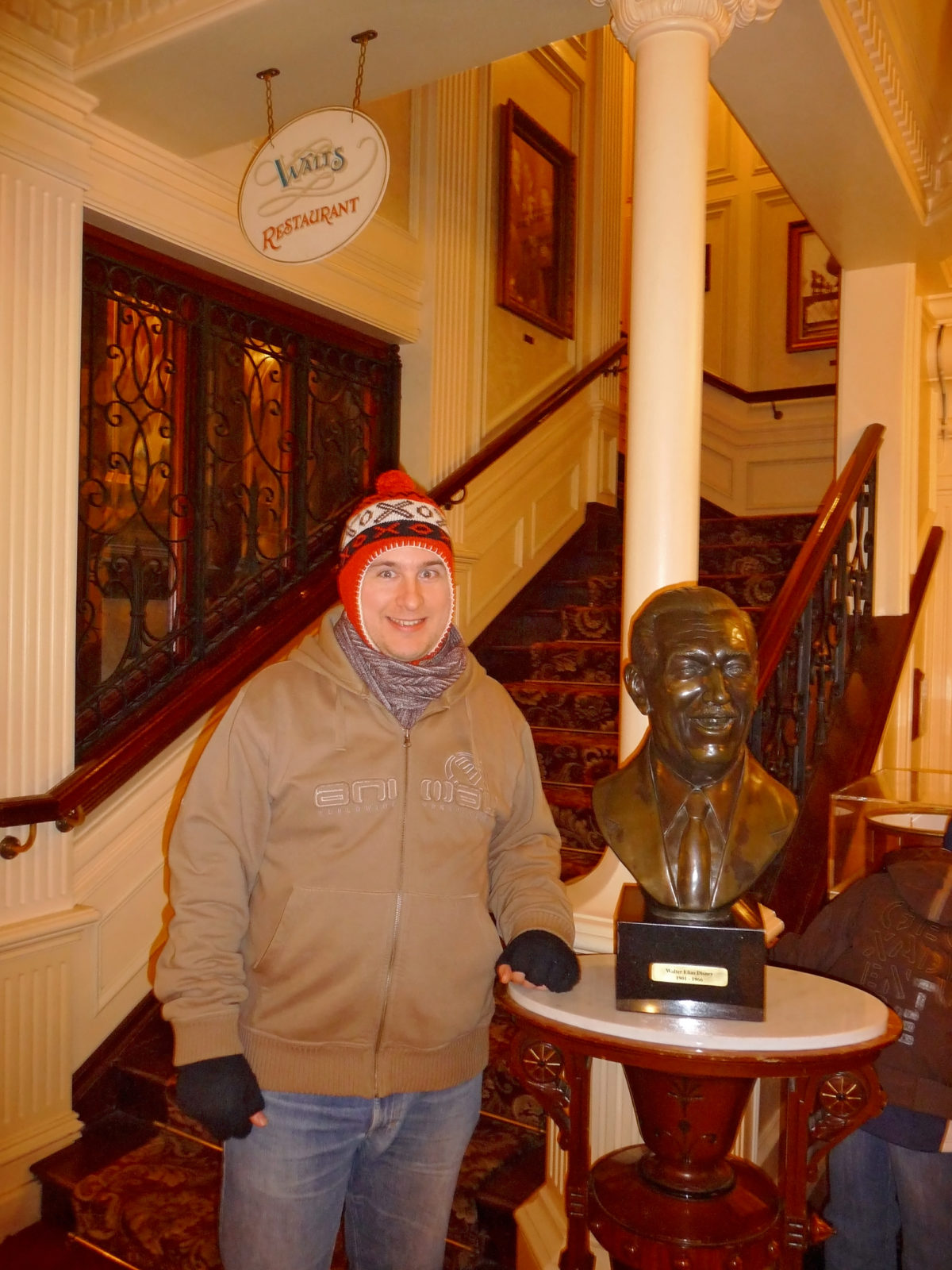 Image shows the bust of walt's head inside Walt's Restaurant in disneyland paris. A man is stood next to it, and he is dressed in warm winter clothes and a hat.
