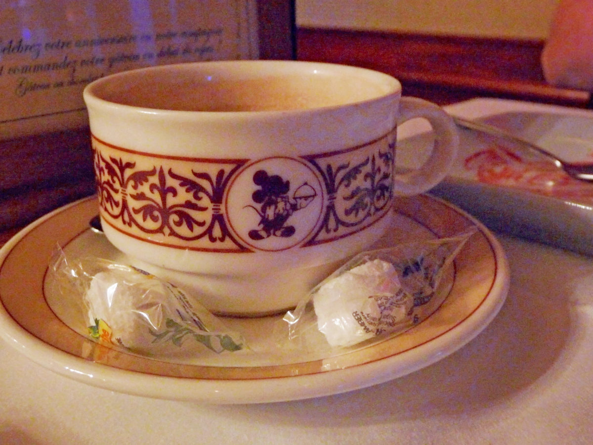 Image shows a coffee cup an saucer with Mickey Mouse as a fine dining server illustrated on the side.  There are two sugar cubes in plastic wrappers on the saucer.