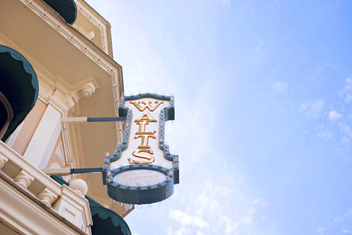 Image shows Walt's pink and green and gold Restaurant Signage on the side of main street building in Disneyland Paris. The sky is blue.