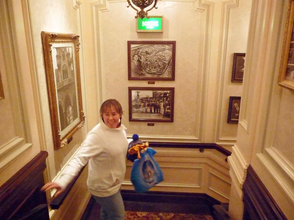 Image shows the interior of the staircase at Walt's restaurant in Disneyland Paris. There are photographs of Disney days gone by on the walls and a women is looking at them as she holds tigger gloves in her hand an a disneyland paris shopping bag.