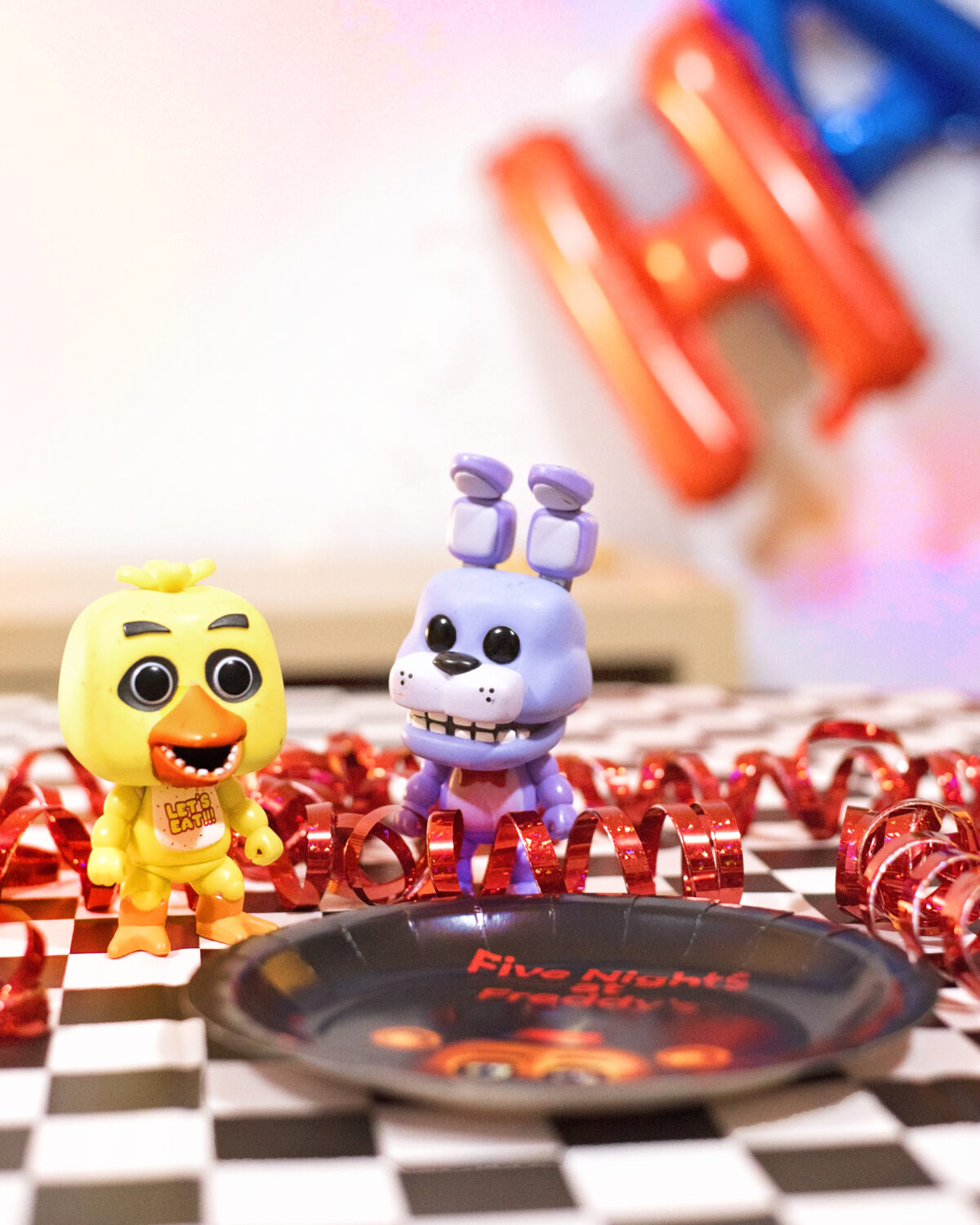 Image shows Party table close up of Pop Vinyl Five Nights at Freddy's figures Bonnie and Chica. They are surrounded by metallic red streamers and there are balloons in the background. Image by keep up with the jones family.