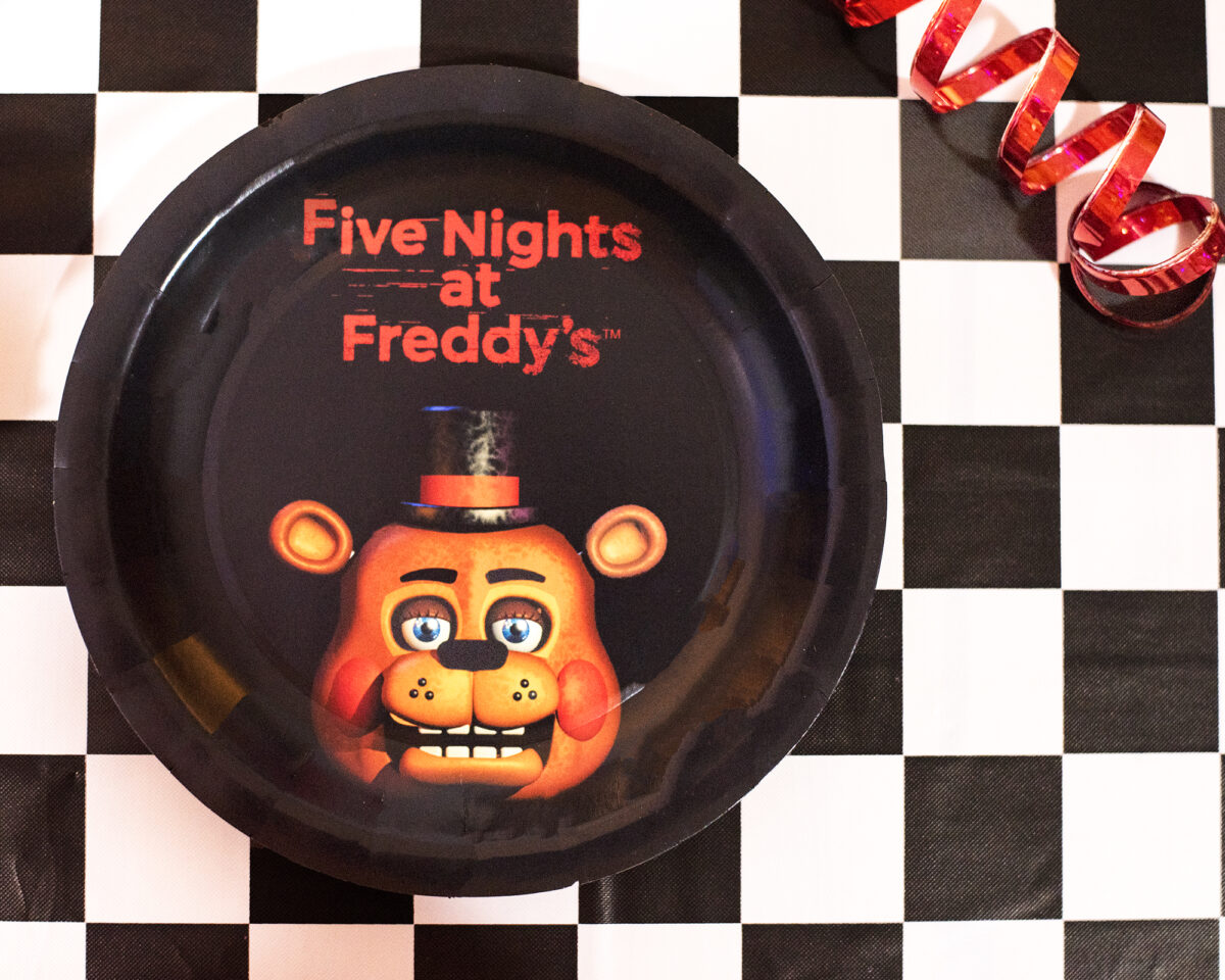 Image shows a checkered black and white tablecloth set with metallic red streamers and a black paper Five NIghts at Freddy's plate featuring Freddy Fazbear's face.  Image by keep up with the jones family.