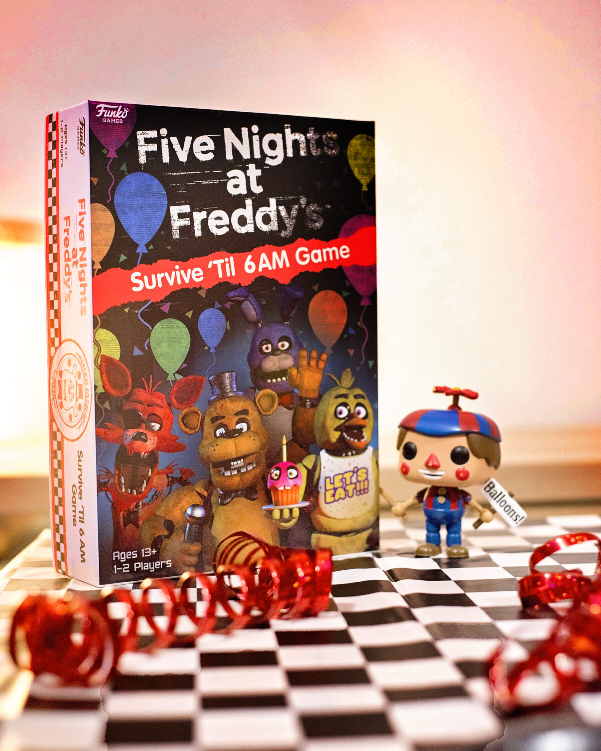Image shows the Five Nights at Freddy's Survive 'til 6AM game on a table covered with a black and white checkered tablecloth, strewn with red metallic streamers and with a Funko Pop Vinyl figure of Balloon boy from FNAF stood next to the box. Image by keep up with the jones family.