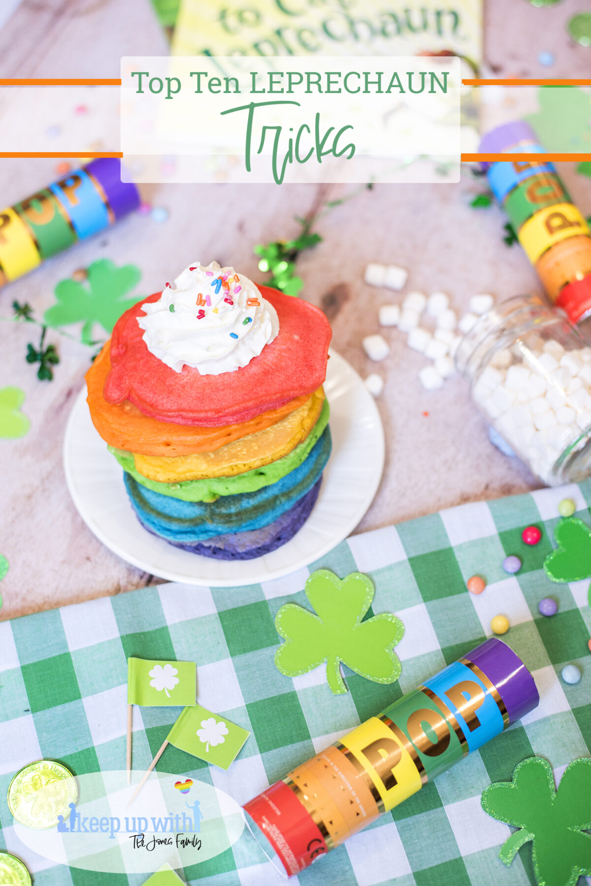 Top ten leprechaun tricks for st. patricks day. Image shows stack of rainbow pancakes on top of a St. Patrick's day tablescape.  Green checkered gingham tablecloth, leprechaun gold and rainbow confetti cannons.  There are little green flags featuring white shamrocks and glittery card shamrocks, rainbow coloured sweets and candies and a jar of spilled marshmallows. 
