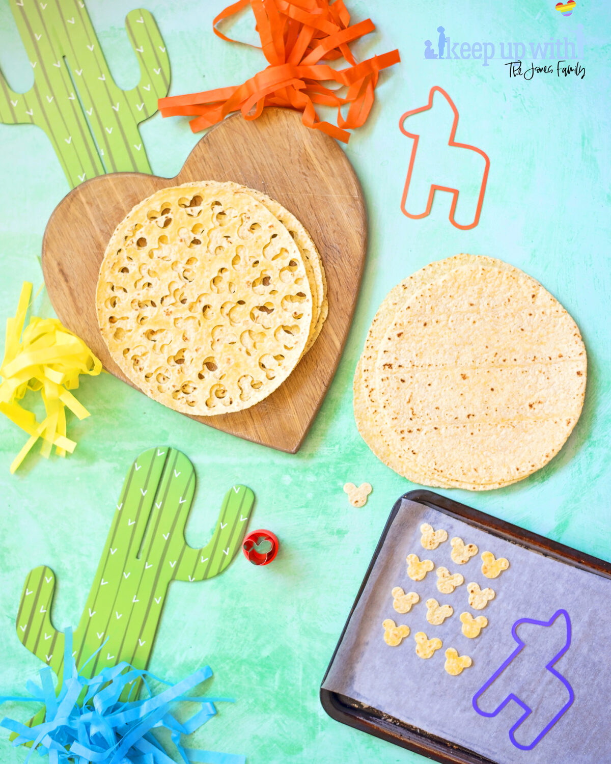 Image shows the recipe for Mickey Mouse taco chips. There is a baking tray with small cut out tortilla chips in the shape of Mickey Mouse's head. There is a metal Mickey Mouse cookie cutter and decorations for cinco de mayo, disney style.  There is a cardboard cactus and some tassles in bright colours on a sea green background.  Keep Up with the Jones Family.