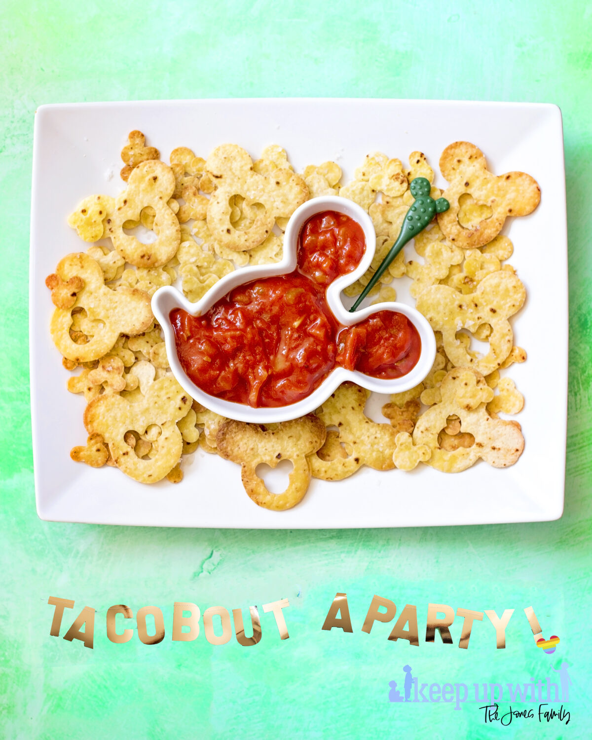 Image shows large rectangular white plate filled with Disney's Mickey Mouse nacho chips and a Mickey Mouse shaped bowl of salsa.  The background is sea green and there is a golden banner across the bottom saying "taco bout a party" for cinco de mayo. Keep up with the jones family.