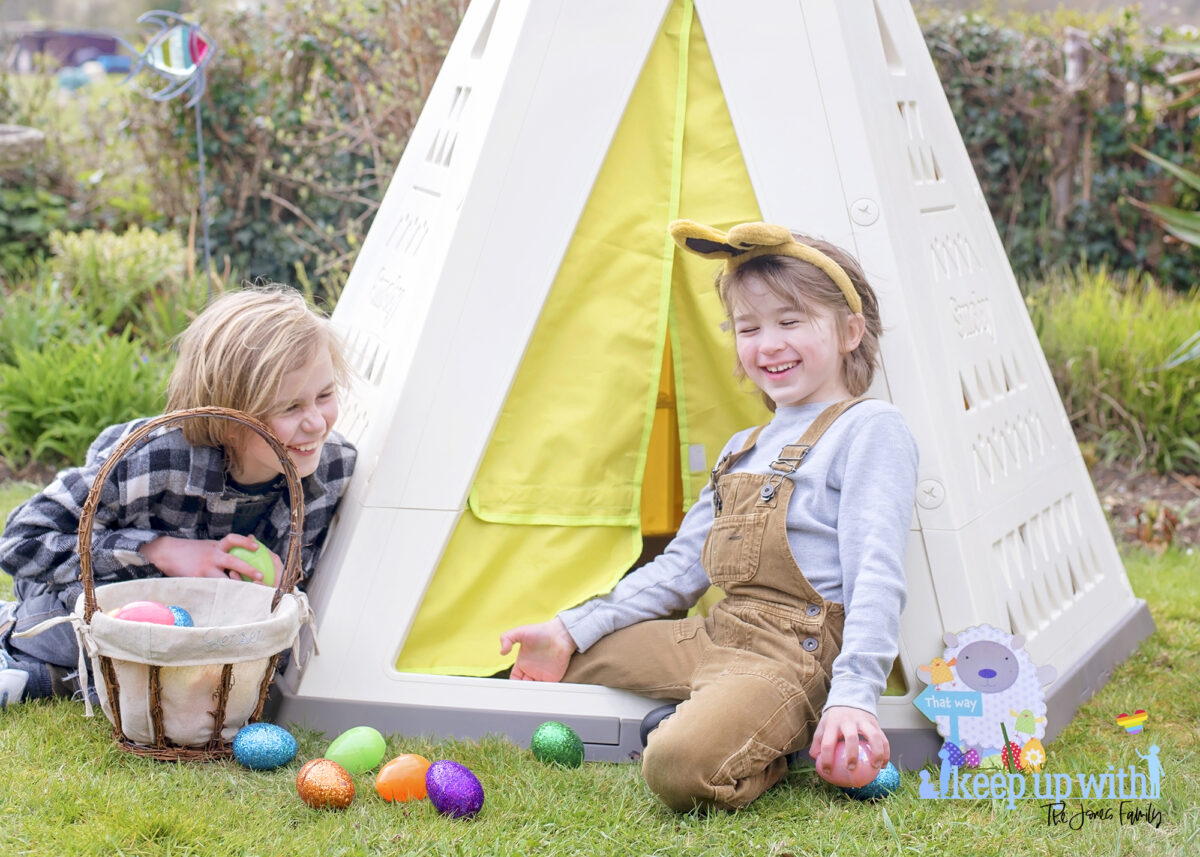 Image shows the new Smoby Teepee, available on Amazon, in the garden. The teepee is cream, white and green with a triangle pattern on the sides.  There is a fabric door which is lime green and two little boys are sat on the outside of the teepee, dressed for easter with bunny ears on.  They are laughing.  They have easter baskets filled with coloured easter eggs which have spilled onto the floor.  The teepee is decorated with a sign which reads Easter Bunny Stop here!