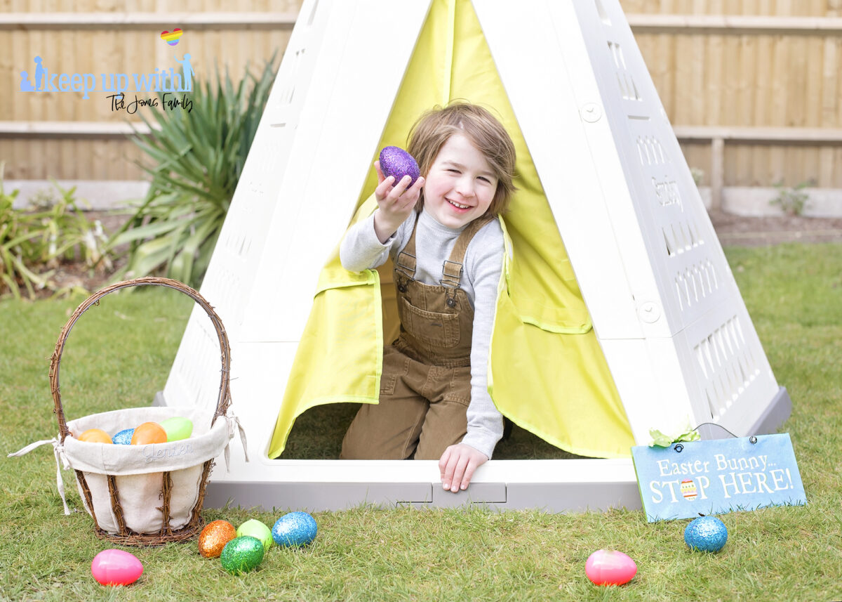 Image shows the new Smoby Teepee, available on Amazon, in the garden. The teepee is cream, white and green with a triangle pattern on the sides.  There is a fabric door which is lime green and a little boy with brown hair is sat in the doorway, dressed for easter and holding a purple glittery easter egg. He is laughing.  There is an easter basket filled with coloured easter eggs which have spilled onto the floor.  The teepee is decorated with a sign which reads Easter Bunny Stop here!