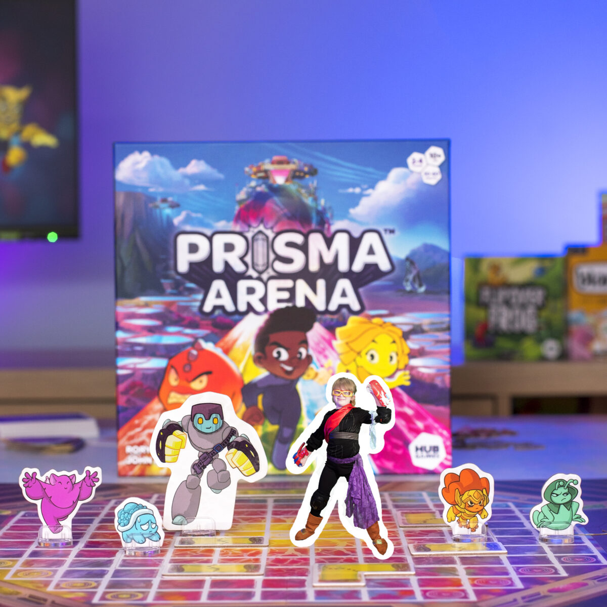 Prisma Arena Tabletop Game from Hub Games. Picture shows Heroes and Mo'kons from the game with a child posing as one of the game standee pieces.  The colours are bright and vivid.