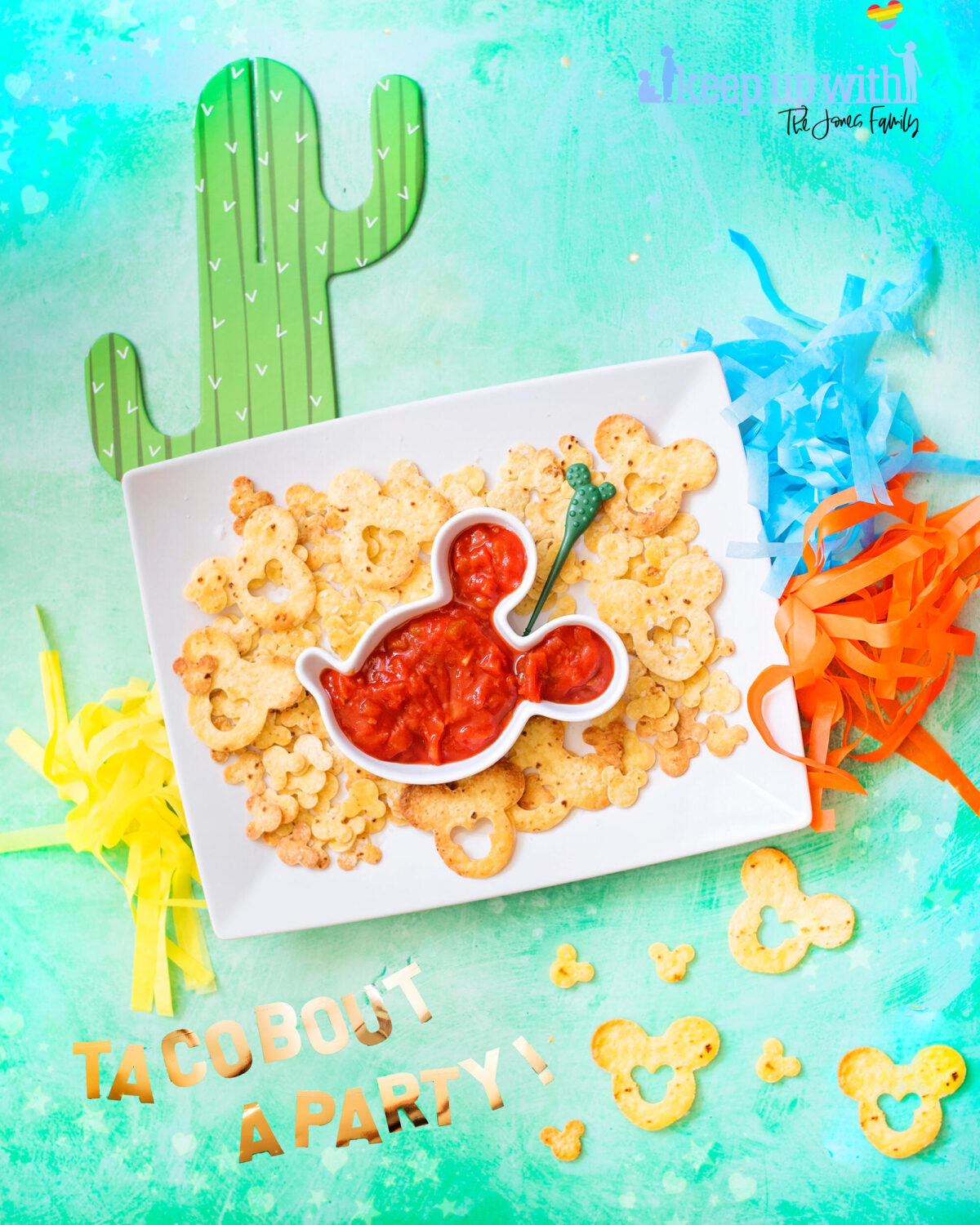 Image shows rectangular white plate filled with Mickey Mouse shaped tortilla chips and a Mickey Mouse head shaped salsa bowl. Surrounding the plate are cinco de mayo disney decorations. There are cardboard cactus and orange, blue and yellow pom pom tassles.  They are Mickey Mouse Nachos, fun food for children 