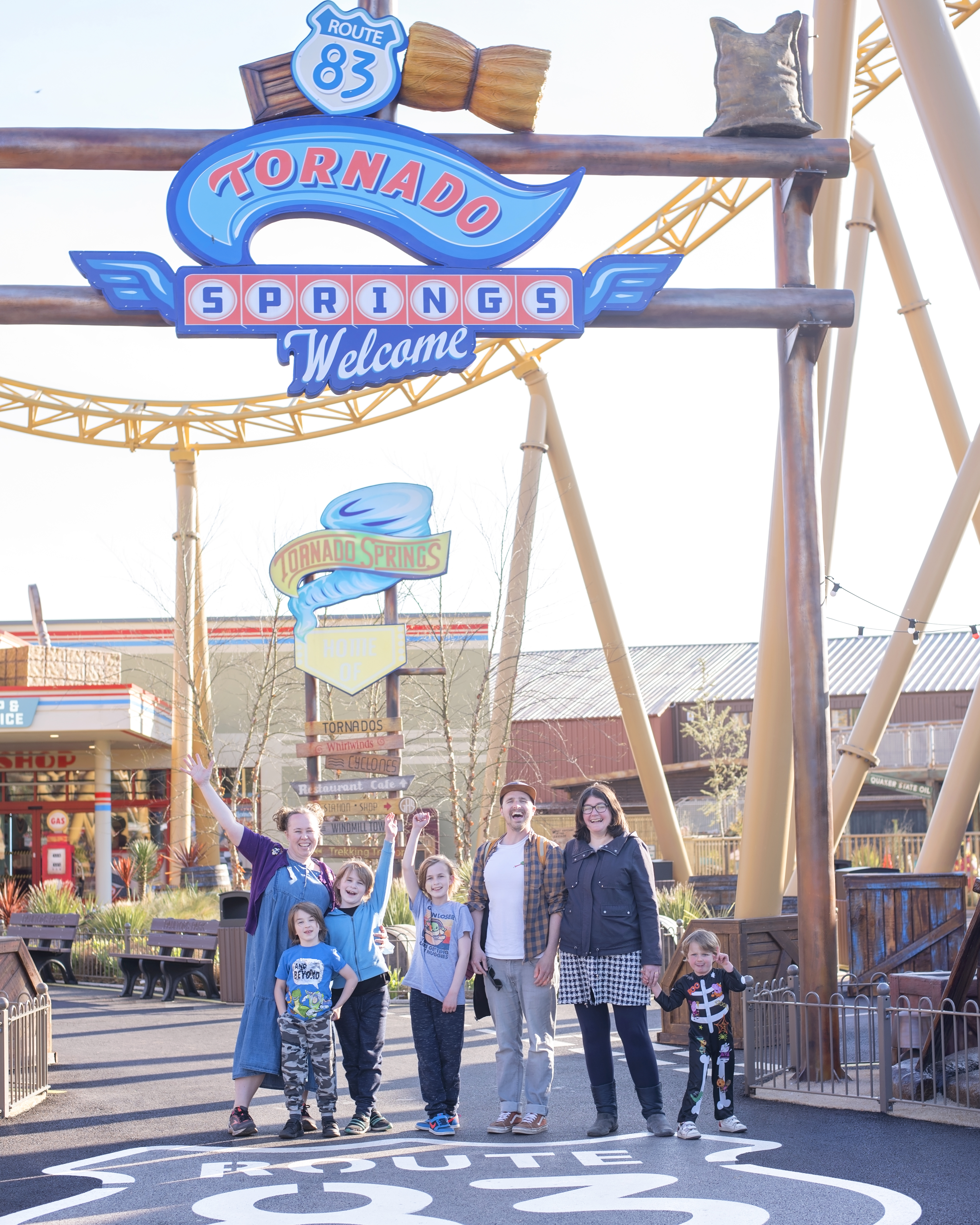 Image shows a family of seven stood underneath the Paultons park Tornado Springs rollercoaster and sign in ower, hampshire. The rollercoaster is the yellow storm chaser new for 2021 and on the floor is a large painted white route 83 logo.