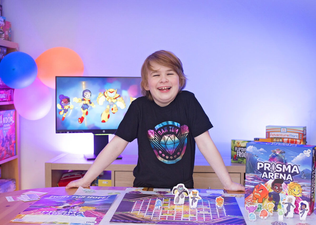 Photo shows a boy stood over a tabletop looking at the Prisma Arena Game set up.  He has blond hair and is smiling.  The boy is wearing a black Dan TDM t-shirt with a colourful foil diamond logo on it. The lighting in the photo is blue and behind the boy is a monitor displaying graphics from the Hug Games website.  Other Hub Games are in the background including Flip Over Frog and Adventure Mart.