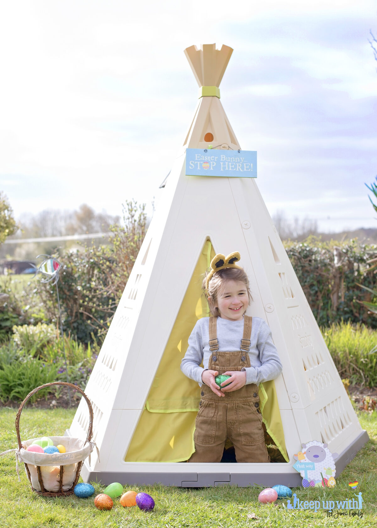 Image shows the new SmobyUK Teepee, available on Amazon, in the garden. The teepee is cream, white and green with a triangle pattern on the sides.  There is a fabric door which is lime green and a little boy aged 6 is stood in the doorway, dressed in light tan dungarees and a pale blue top, and wearing grey and pink bunny ears for easter.   He is laughing.  There is an easter baskets filled with coloured easter eggs which have spilled onto the floor.  The teepee is decorated with a sign which reads Easter Bunny Stop here!