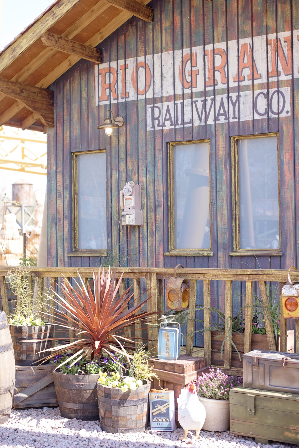 Image shows theming at Paultons Park Tornado Springs area. An american wooden building belonging to the rio grande railway company. There is a white chicken walking on the porch, with flowers in barrels and cans of aviation oil littered next to ammunition cases. 