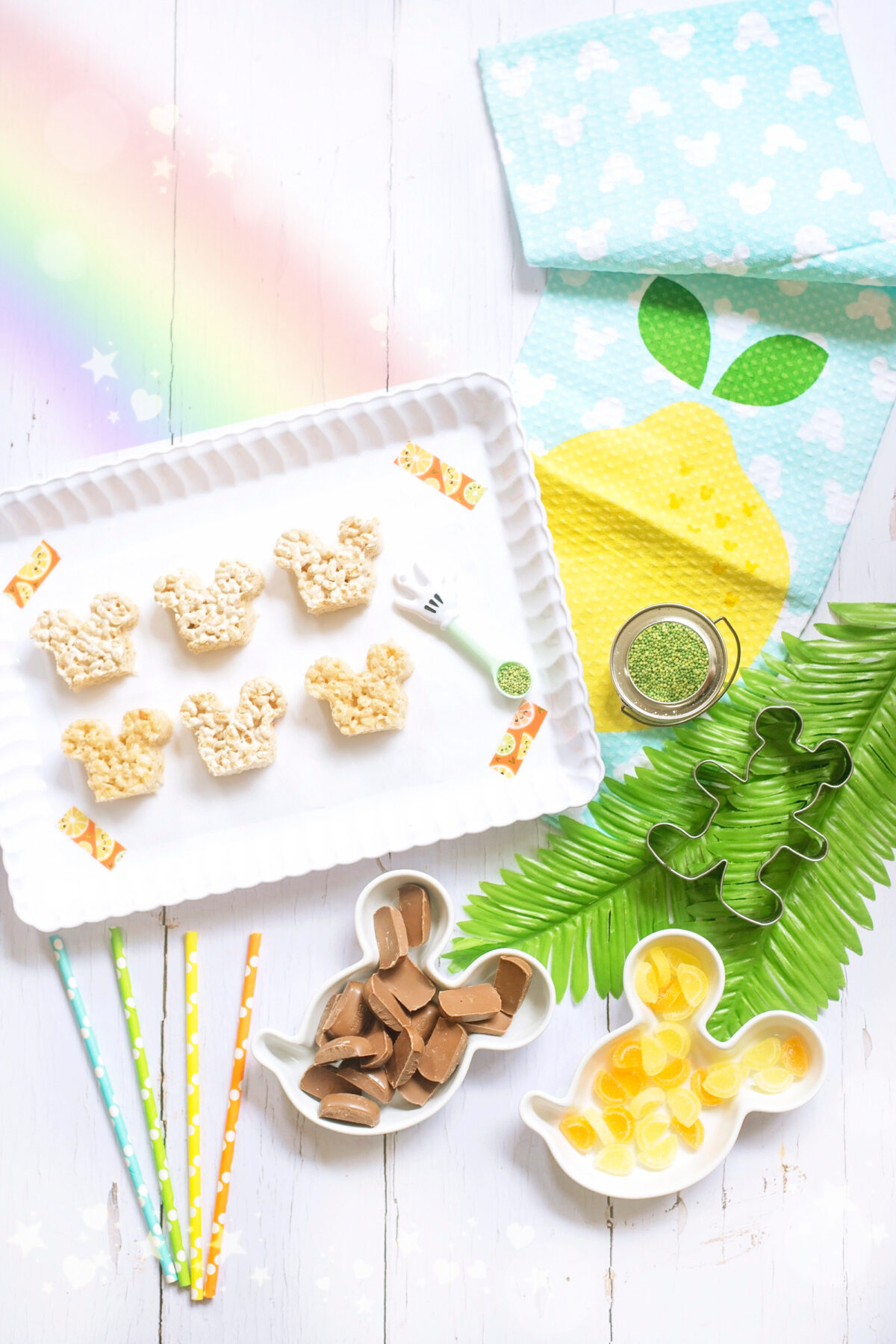 Image shows scalloped tray of summer themed Mickey Mouse Rice Krispie Treats surrounded by orange and lemon slices and tropical leaves, Disney tea towels and Minnie Mouse straws. All of the ingredients for the recipe are laid out on the tray including a DIsney's Mickey Mouse cookie cutter.