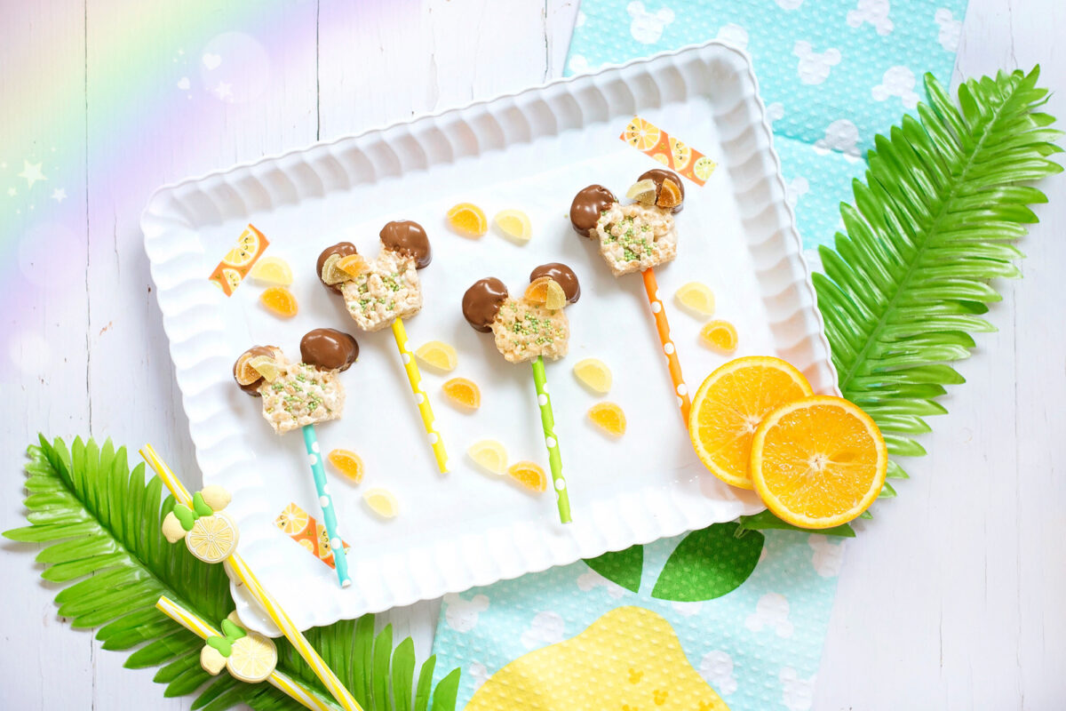 Image shows scalloped tray of summer themed Mickey Mouse Rice Krispie Treats surrounded by orange and lemon slices and tropical leaces, Disney tea towels and Minnie Mouse straws.