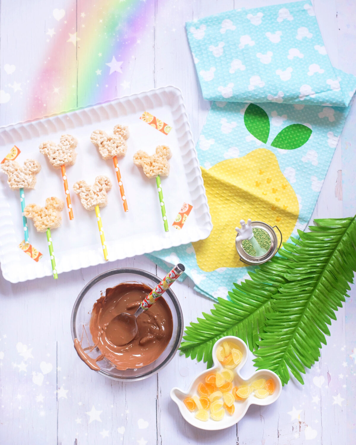 Image shows scalloped tray of summer themed Mickey Mouse Rice Krispie Treats surrounded by orange and lemon slices and tropical leaves, Disney tea towels and Minnie Mouse straws. There is a bowl of melted chocolate ready to dip the Mickey Mouse treats into.