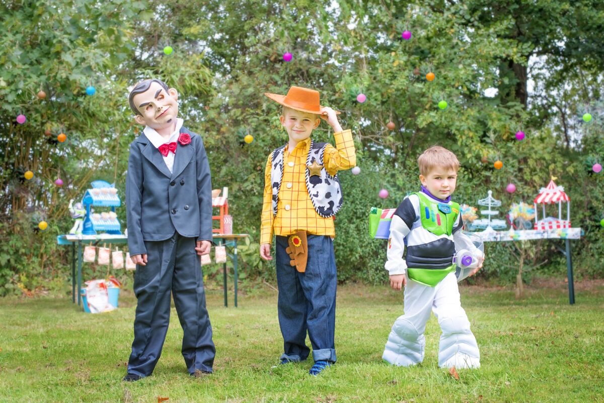 Image shows three of the Jones boys standing in their party dress up clothes ready for the Disney Toy Story Birthday party. One is Buzz Lightyear, one is Woody the Cowboy, and the third is dressed like the ventriloquist's dummy from Toy Story 4 who help Gabby.  They are outside in the garden and there are party tables behind them.