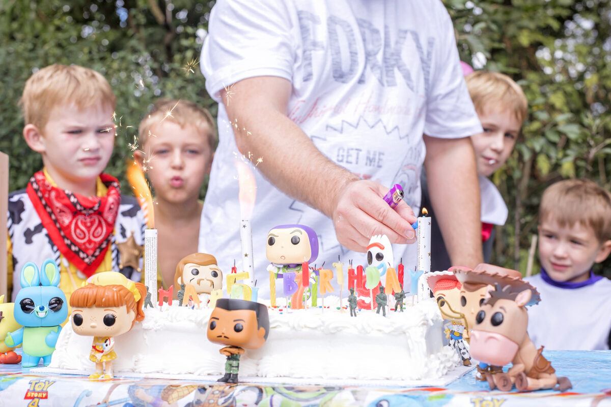 Image shows a large white Costco cake covered in Toy Story 4 Pop Funko toys.  Combat Carl, Woody, Bullseye, Gabby, Bunny and Ducky, Buzz, Jessie, Forky and Duke kaboom can be seen.  In the background there is an adult lighting the candles on the cake and the children of the jones family are watching.
