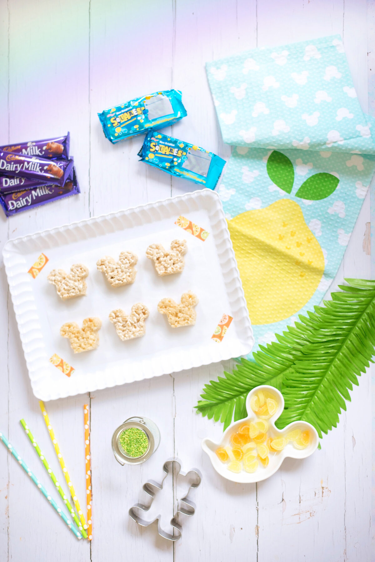 Image shows all of the ingredients needed to make the summer themed Disney's Mickey Mouse Rice Krispie Treats. Dairy Milk chocolate bars, rice krispie squares, green sprinkles, orange and lemon slices.  They are laid ona white wooden backdrop with a Mickey Mouse lemon tea towel and a white scalloped tray, with a Mickey Mouse cookie cutter and coloured spotted paper straws.