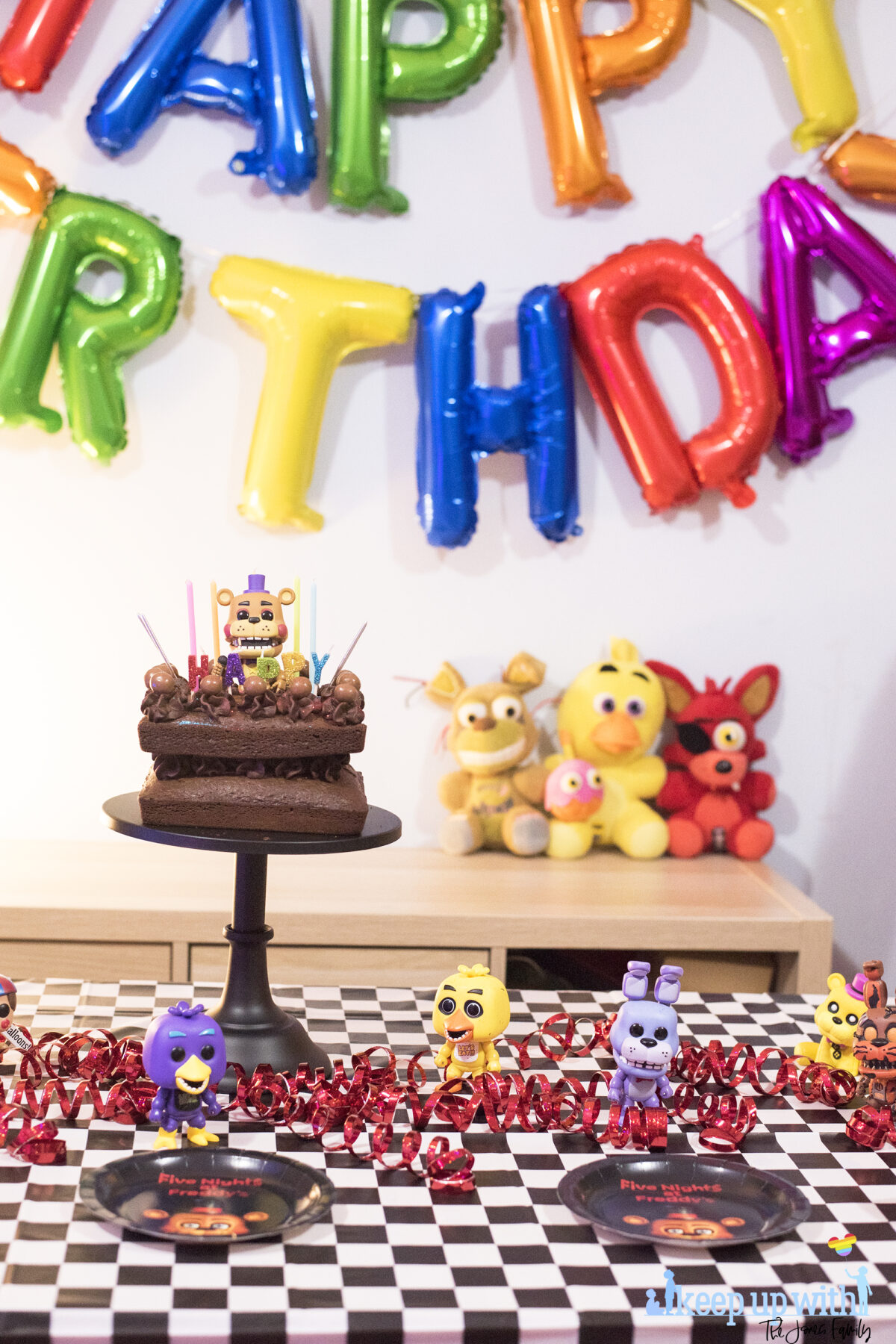 How to throw a Five Nights at Freddy's Party. Image shows a party table covered in a black checkered tablecloth, happy birthday spelled out in rainbow coloured balloons as a garland across the wall, three FNAF plushies; Foxy, Chica and Plushtrap sat on the sideboard and a chocolate cake on a black cake pedestal with a Funko Pop Viny; Rockstar Freddy on top of it. Image by Keep Up With The Jones Family