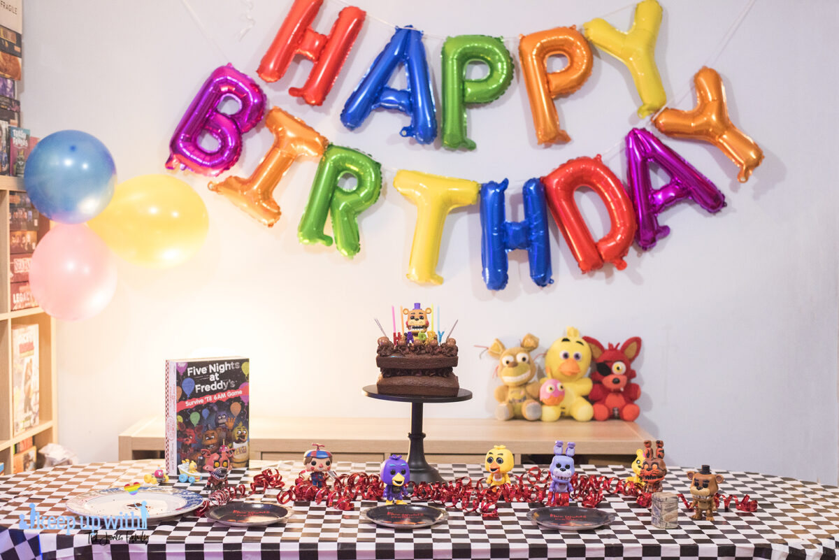 How to throw a Five Nights at Freddy's Party. Image shows a party table covered in a black checkered tablecloth, happy birthday spelled out in rainbow coloured balloons as a garland across the wall, three FNAF plushies; Foxy, Chica and Plushtrap sat on the sideboard and a chocolate cake on a black cake pedestal with a Funko Pop Vinyl Rockstar Freddy on top of it. The FNAF board game is on the table with more Funko figures - chica, jack-o-bonnie, black light chica and balloon boy.
