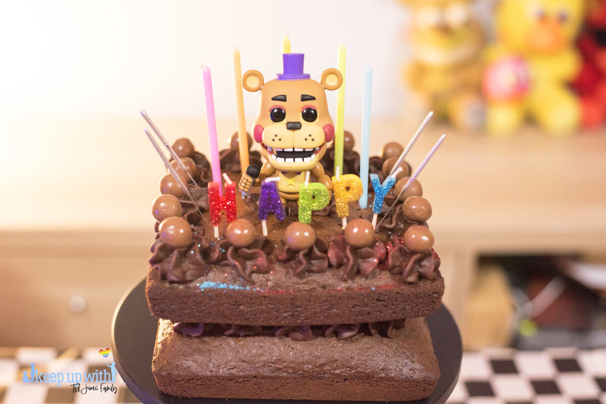 Image shows a homemade square chocolate cake decorated with candles in different colours saying "happy:. There are chocolate icing swirls set with maltesers on top and there are sparklers ready to be lit sticking out of the cake.  In the middle of the cake sits a Funko Pop Vinyl sigure of Freddy Fazbear.