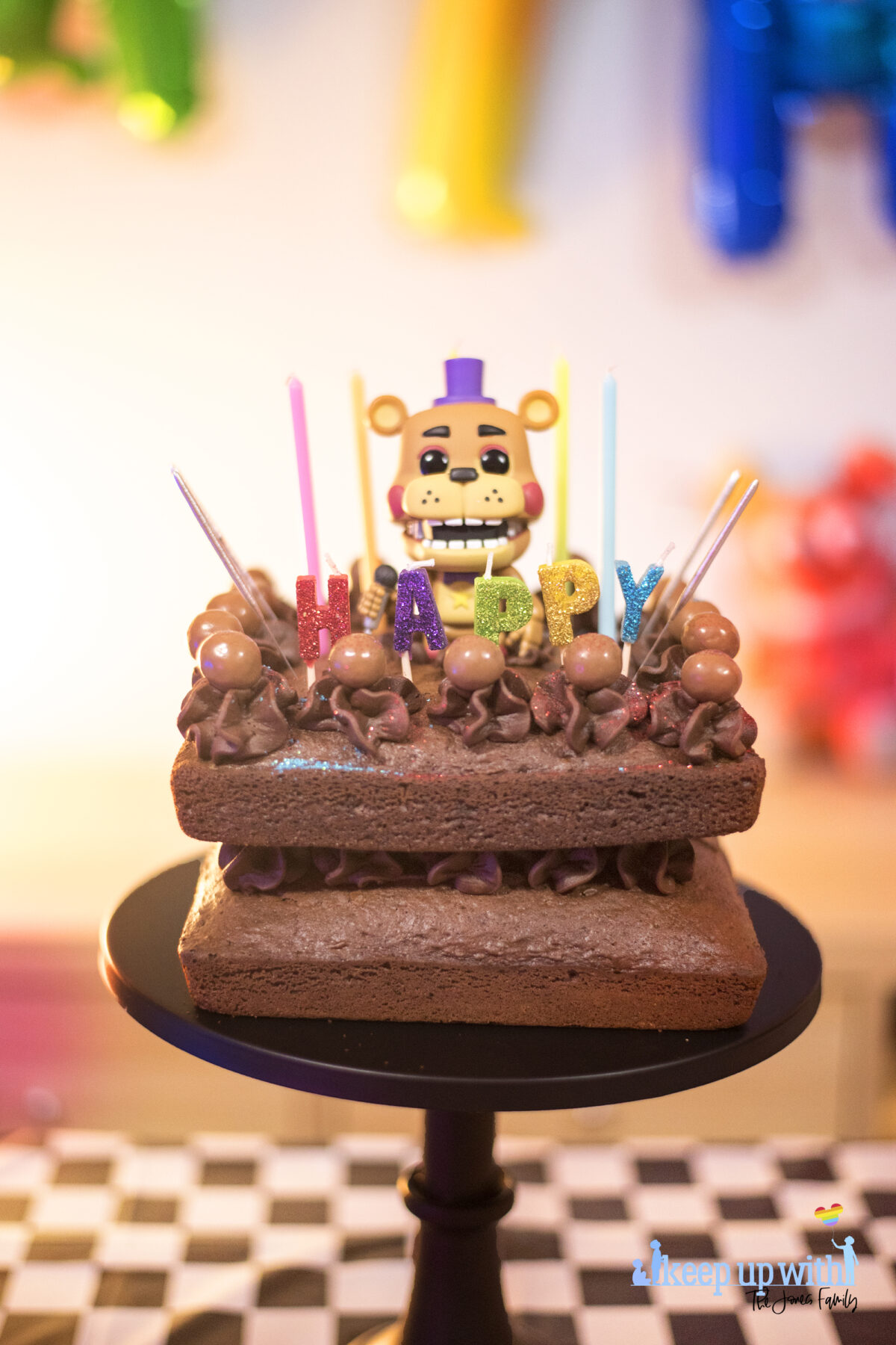 Image shows a homemade square chocolate cake decorated with candles in different colours saying "happy:. There are chocolate icing swirls set with maltesers on top and there are sparklers ready to be lit sticking out of the cake.  In the middle of the cake sits a Funko Pop Vinyl sigure of Freddy Fazbear. Image by keep up with the jones family.