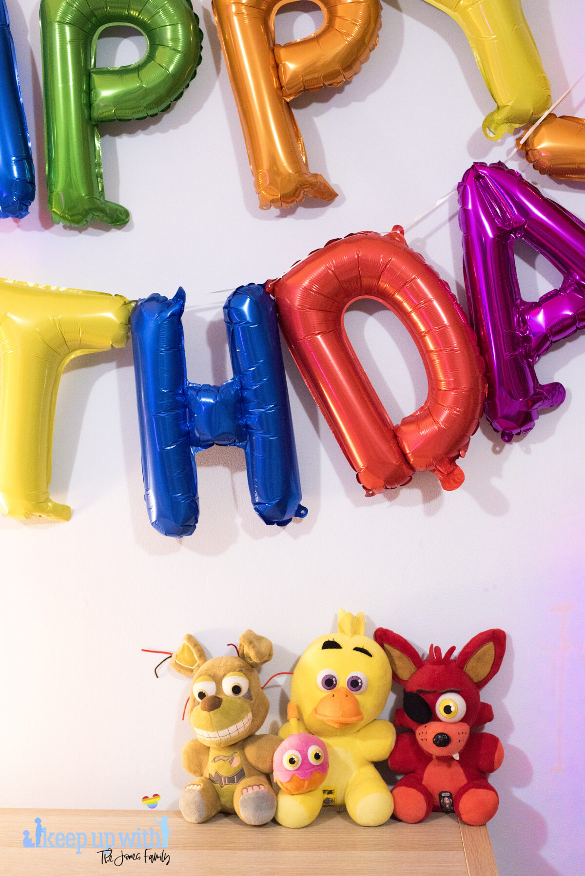 Image shows three Five Nights at Freddy's large plush toys. Foxy, Chica and Springtrap. They are sat on a wooden sideboard against a pale blue wall and the coloured happy birthday balloons are on the wall in the background. Image by keep up with the jones family.