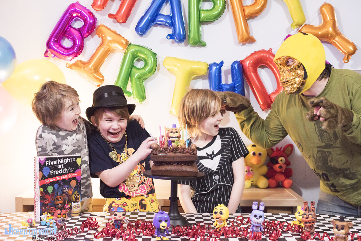 Image shows three boys enjoying the Five Nights at Freddy's Party table. There are Pop Vinyl Funko FNAF figures, a chocolate cake on a black pedestal and the Five Nights at Freddy's Board game, Survive 'til 6am.  A man dressed up with a metallic skull mask and wolf paw gloves is pretending to scare them from the right hand side of the image. Image by Keep up with the jones family.