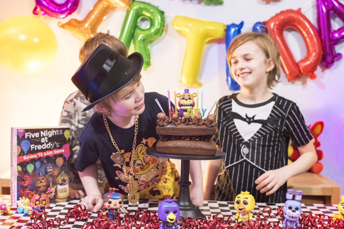 Image shows three boys enjoying the Five Nights at Freddy's Party table. There are Pop Vinyl Funko FNAF figures, a chocolate cake on a black pedestal and the Five Nights at Freddy's Board game, Survice 'til 6am. Image by Keep up with the jones family.
