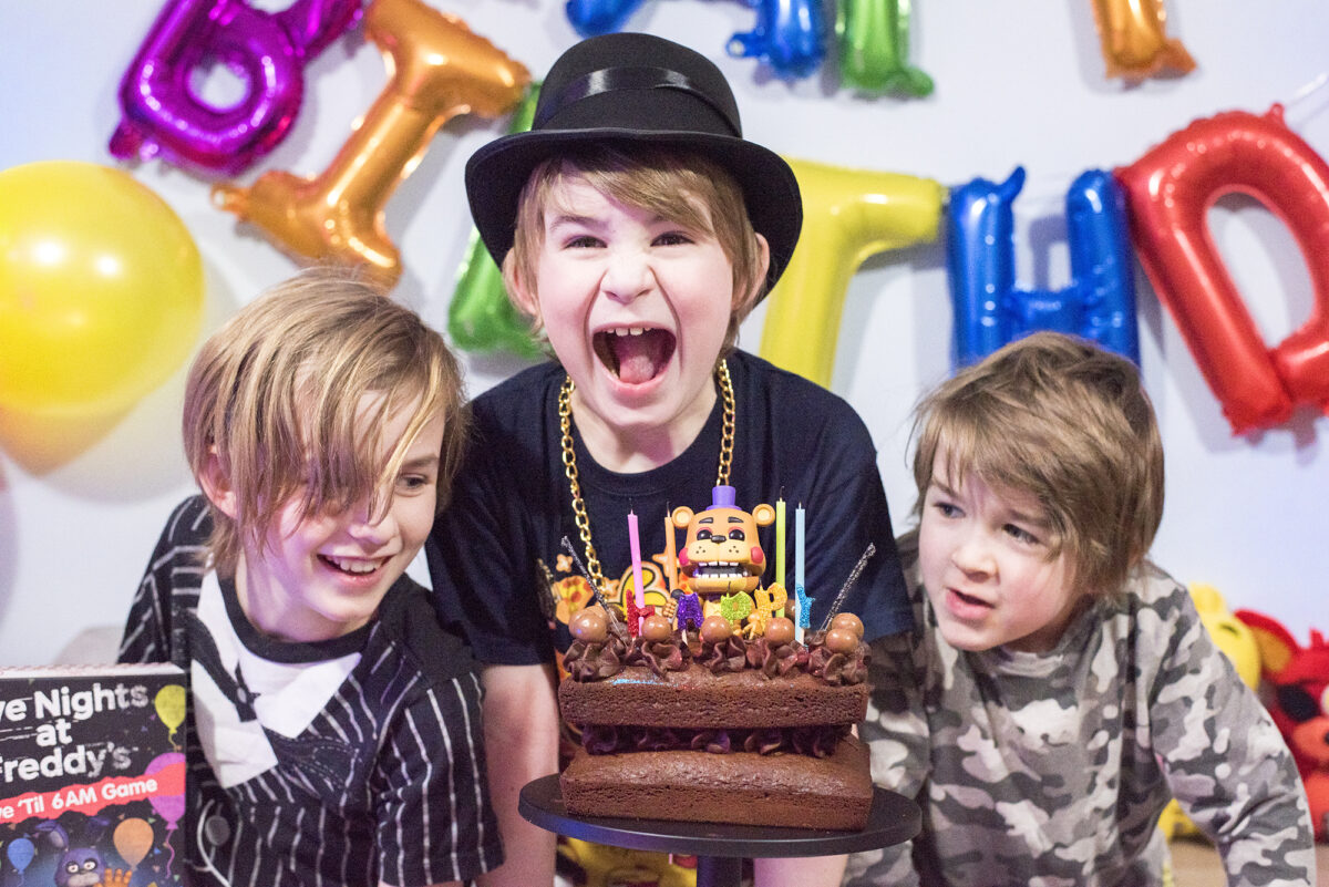 Image shows three boys enjoying the Five Nights at Freddy's Party table. There are Pop Vinyl Funko FNAF figures, a chocolate cake on a black pedestal and the Five Nights at Freddy's Board game, Survive 'til 6am. Image by Keep up with the jones family. The middle boy is pretending to take a bit bite of the cake and the other two are smiling.