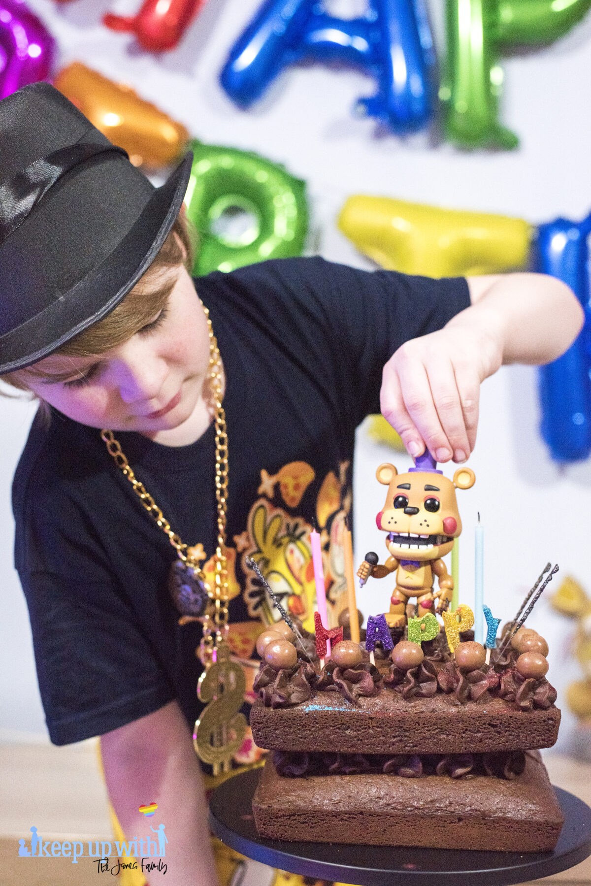 Image shows a blond haired boy wearing a black FNAF chica t-shirt and black top hat with a gold chain. 
 He is peering over a chocolate cake. He is lifting a Freddy Fazbear's Funko Pop Vinyl figure from Five Nights at Freddy's off the cake.