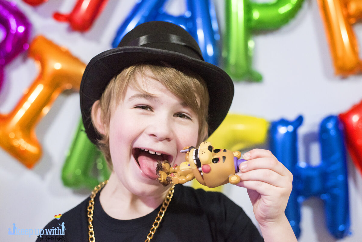 Image shows a blond haired boy wearing a black FNAF chica t-shirt and black top hat with a gold chain. 
 He is pretending to lick the chocolate cake off Freddy Fazbear's Funko Pop Vinyl figure from Five Nights at Freddy's which is covered in chocolate frosting from the cake. Image by keep up with the jones family.