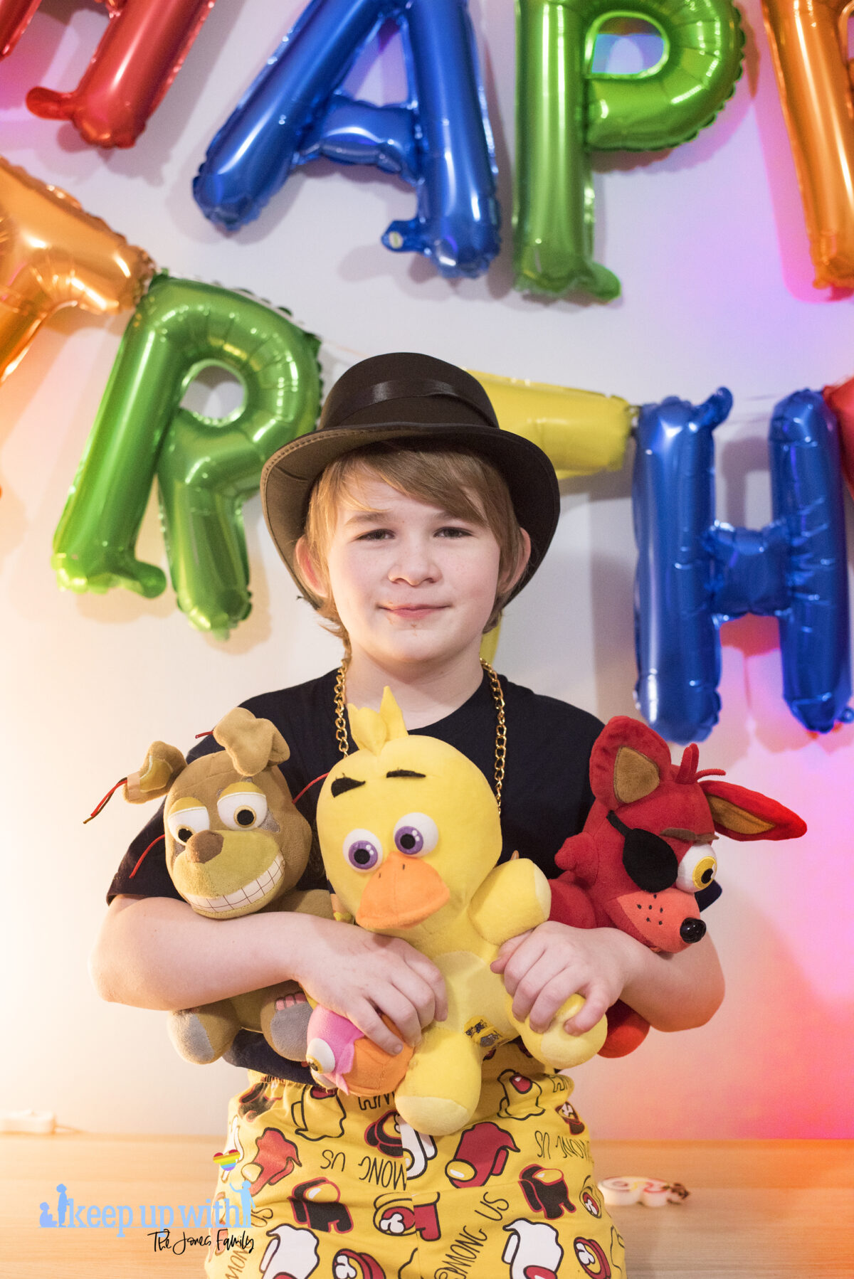 Image shows a blond haired boy with a black tshirt and top hat like Henry Stickman stood with three Five Nights at Freddy's large plush toys. Foxy, Chica and Springtrap. They are sat on a wooden sideboard against a pale blue wall and the coloured happy birthday balloons are on the wall in the background. Image by keep up with the jones family.