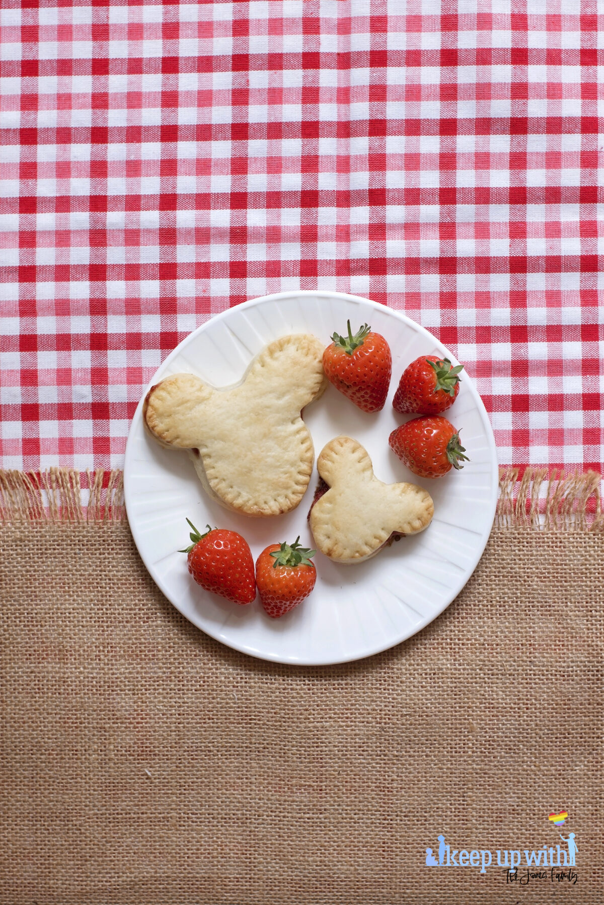 Image shows a flatlay of a red gingham checked tablecloth, overlaid with a jute burlap material with a fringe. On top is a white Vera Wang sideplate with two Mickey Mouse shaped shortcrust pies on. One is larger than the other, and there are strawberries next to the pies. There is a ShopDisney fork balancing on the plate with a summer citrus and apples pattern on it.  The image is watermarked by Keep up with the Jones Family. 