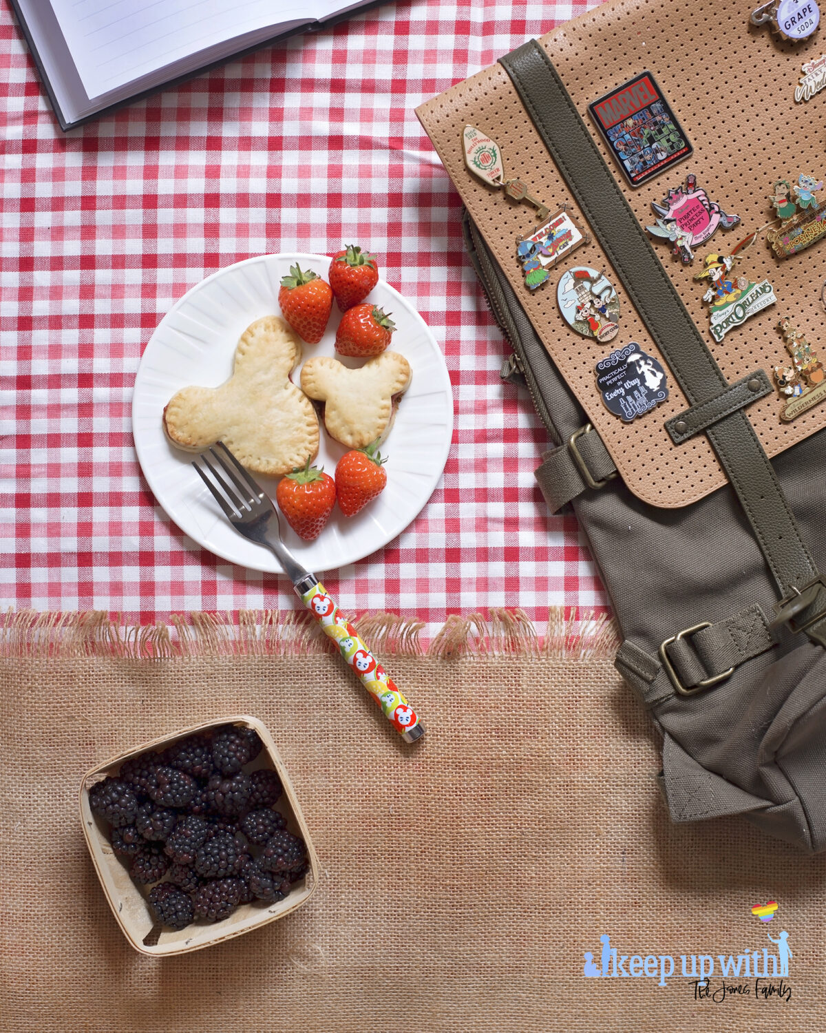 Image shows a flatlay of a red gingham checked tablecloth, overlaid with a jute burlap material with a fringe. On top is a ShopDisneyUK rucksack with many Disney pins from Disney's fairytale weddings, Port Orleans, Castaway Cay, Disneyland Hotel, Cinderellas Table and many more. Next to the bag is a white Vera Wang sideplate with two Mickey Mouse shaped shortcrust pies on. One is larger than the other. There is a ShopDisney fork balancing on the plate with a summer citrus and apples pattern on it.  There is a wooden punnet of blackberries below the plate.The image is watermarked by Keep up with the Jones Family.  