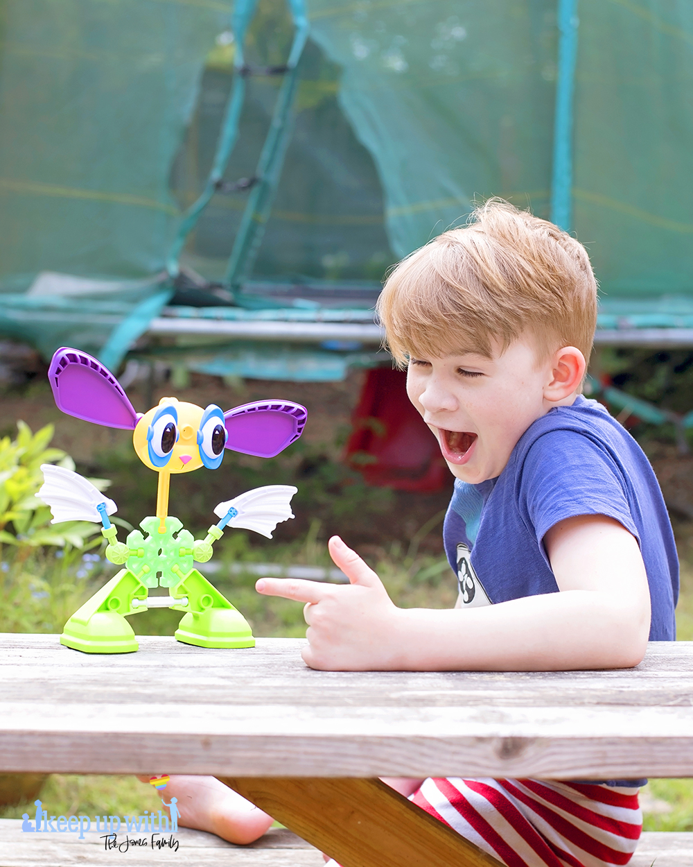 Image shows a boy with blond hair sat at a picnic table, pointing at a toy in front of him.  On the wooden picnic table in front of them is a rabbit like creature, which is made from pieces of the Kid K'nex Budding Builders construction toys box from Basic Fun UK.  By Keep Up With The Jones Family.