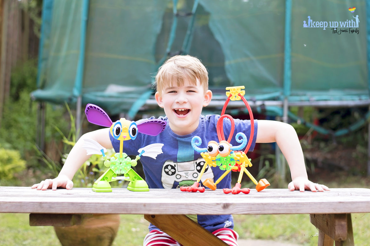 Image shows a boy smiling with his arms on a wooden picnic table in the garden.  On the wooden picnic table in front of him are two creatures which are made from pieces of the Kid K'nex Budding Builders construction toys box from Basic Fun UK.  By Keep Up With The Jones Family.