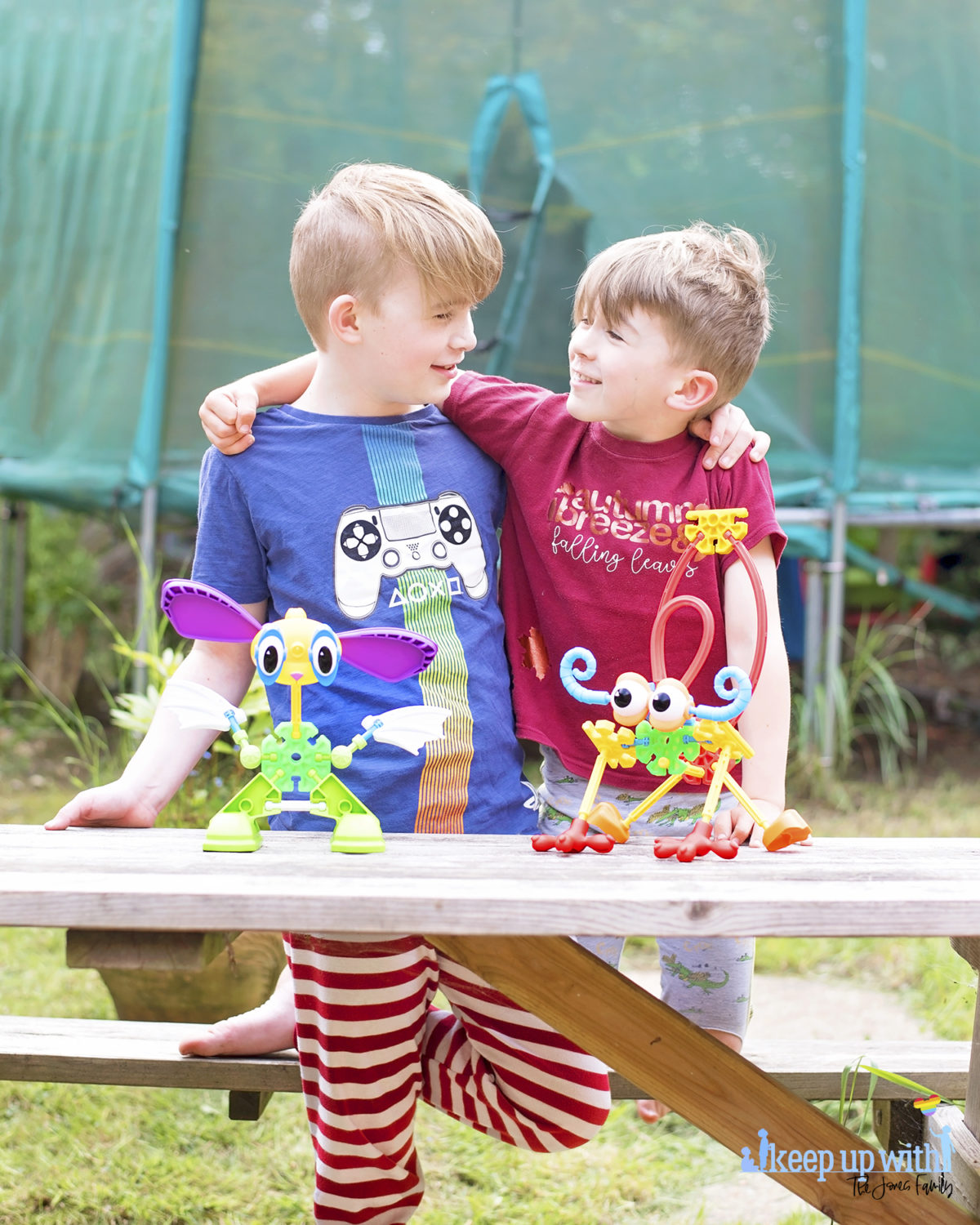 Image shows two boys with their arm around each other's shoulders, looking at each other.  On the wooden picnic table in front of them are two creatures which are made from pieces of the Kid K'nex Budding Builders construction toys box from Basic Fun UK.  By Keep Up With The Jones Family.
