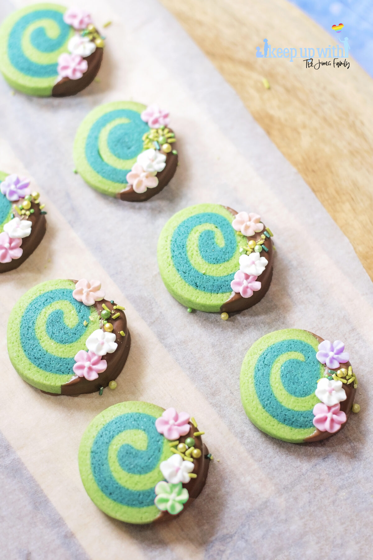 Image shows Disney's Moana Heart of Te Fiti Biscuits, a swirl of bright and dark green, dipped slightly in milk chocolate and embellished with sugar blossom flowers and green sprinkles.  They are set on a light wood chopping board with baking paper underneath. . Image by keep up with the jones family.