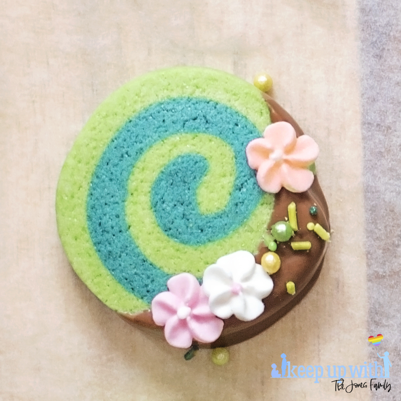 Image shows one of Disney's Moana Heart of Te Fiti Biscuits, a swirl of bright and dark green cookie, dipped slightly in milk chocolate and embellished with sugar blossom flowers and green sprinkles.  They are set on a light wood chopping board with baking paper underneath. . Image by keep up with the jones family.