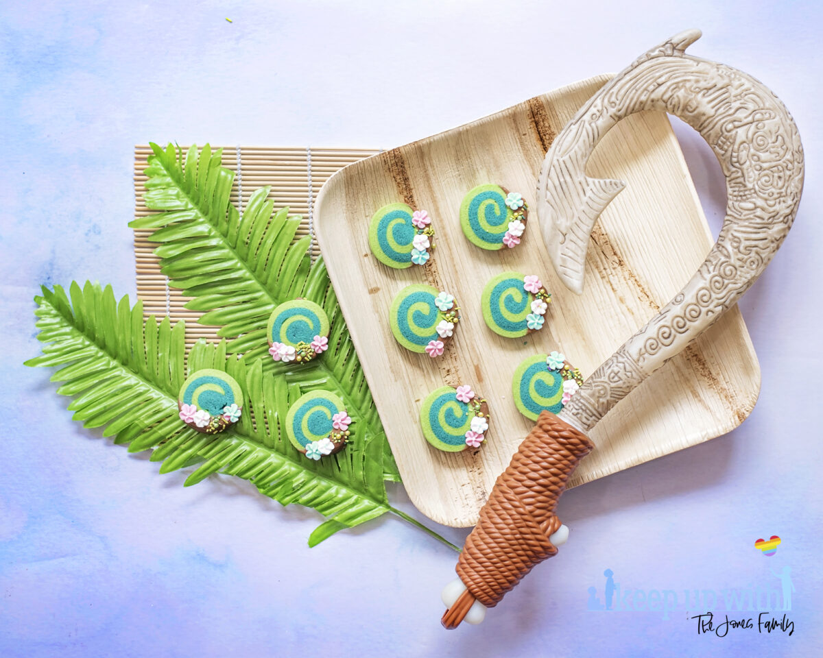 Image shows Disney's Moana Heart of Te Fiti Biscuits, a swirl of bright and dark green cookie, dipped slightly in milk chocolate and embellished with sugar blossom flowers and green sprinkles.  They are set on a bamboo plate with a fern underneath, and a bamboo sushi rolling mat is laid underneath. Maui's fish hook is resting on the plate also. Image by keep up with the jones family.