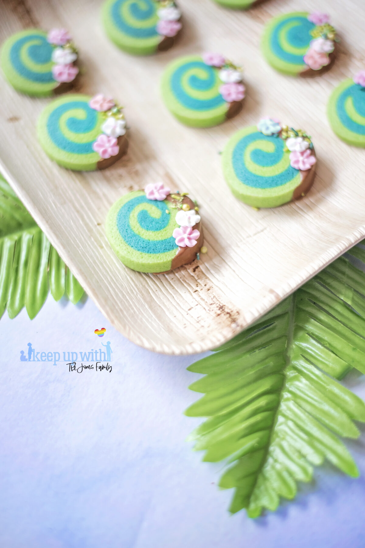 Image shows how to make Disney's Moana Heart of Te Fiti Biscuits, a swirl of bright and dark green, dipped slightly in milk chocolate and embellished with sugar blossom flowers and green sprinkles.  They are set on a bamboo plate with a fern underneath.  Image by keep up with the jones family.