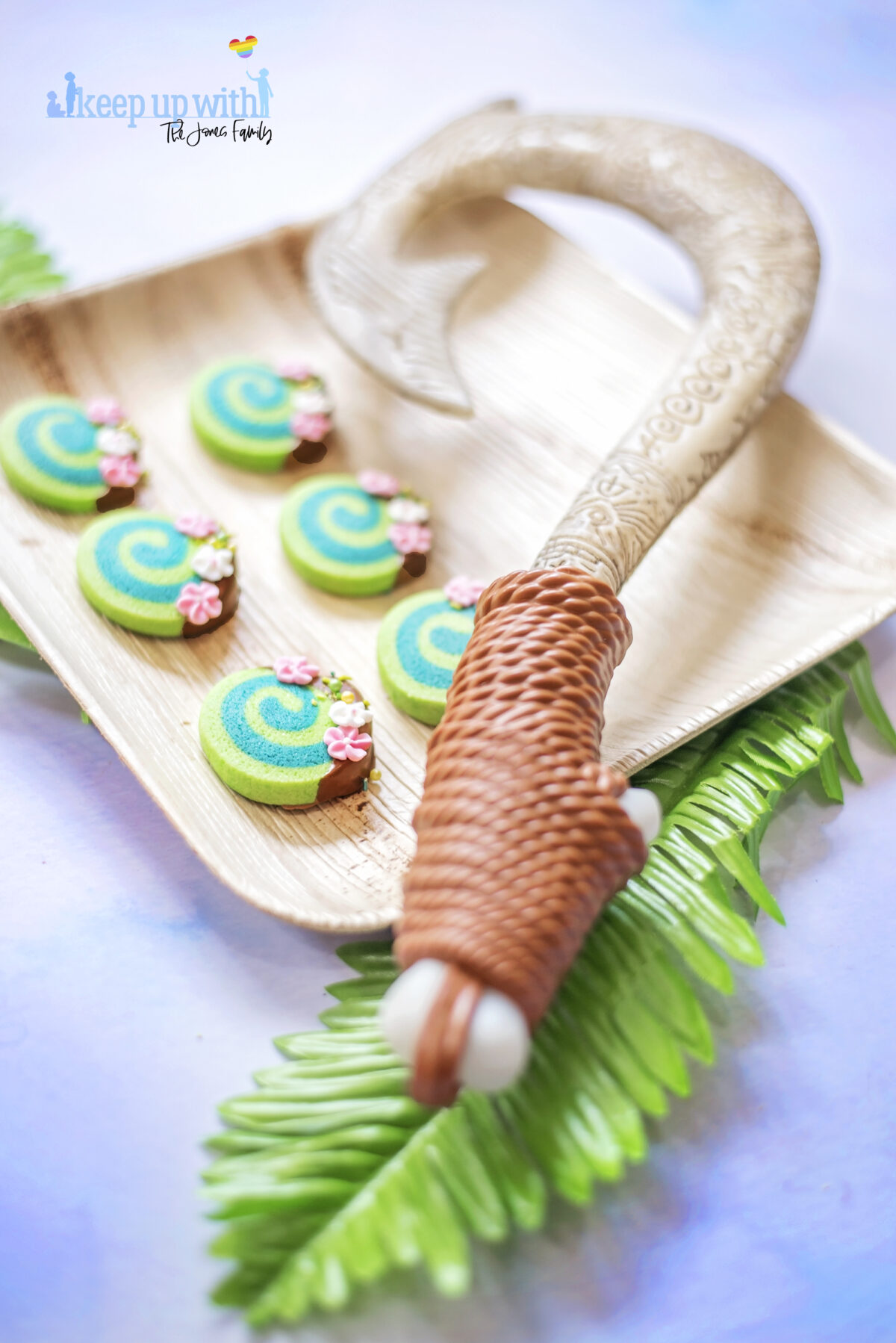 Image shows Disney's Moana Heart of Te Fiti Biscuits, a swirl of bright and dark green, dipped slightly in milk chocolate and embellished with sugar blossom flowers and green sprinkles. They are set on a bamboo plate with a fern underneath, and Maui's fish hook is resting on the plate also. Image by keep up with the jones family.