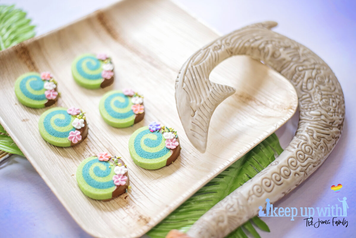 Image shows how to make Disney's Moana Heart of Te Fiti Biscuits, a swirl of bright and dark green, dipped slightly in milk chocolate and embellished with sugar blossom flowers and green sprinkles.  They are set on a bamboo plate with a fern underneath, and Maui's fish hook is resting on the plate also. Image by keep up with the jones family.
