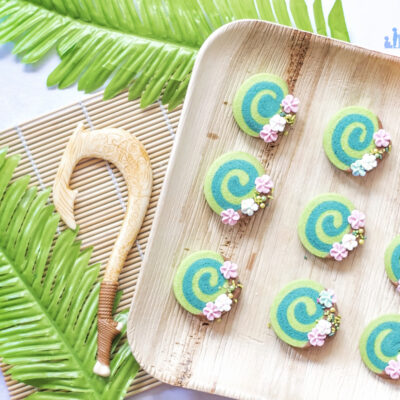 Image shows how to make Disney's Moana Heart of Te Fiti Biscuits, a swirl of bright and dark green, dipped slightly in milk chocolate and embellished with sugar blossom flowers and green sprinkles. They are set on a bamboo plate with a fern underneath, and a small toy version of Maui's fish hook is resting next to the plate also. Image by keep up with the jones family.