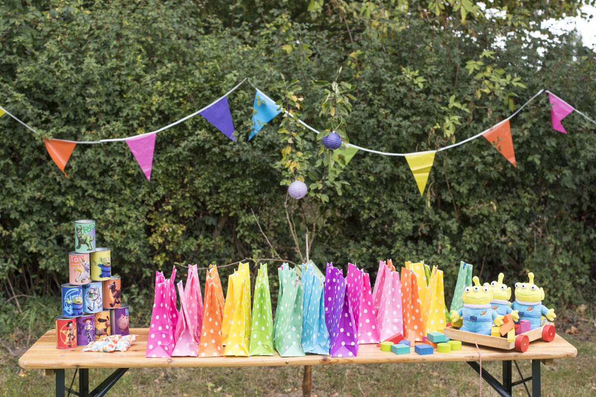 Image shows a Toy Story Party table outdoors with rainbow coloured bunting in the background.  The tabletop is filled with bright coloured, spotted party bags and there are three Toy Story plush Alien toys and a set of knock down cans on the table too. 