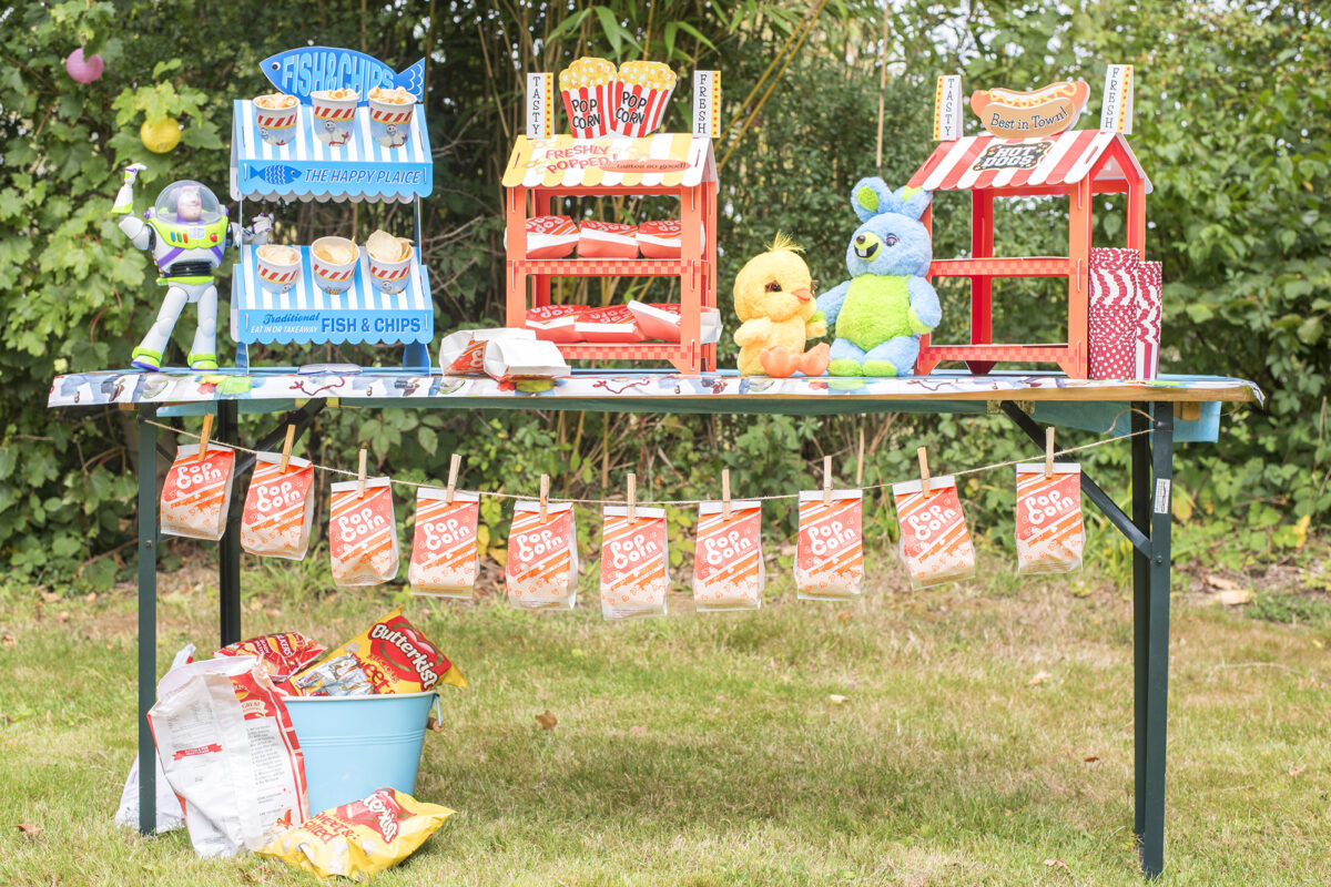 Image shows a party table outdoors in a field. The table has three novelty food stands on top; fish and chips, popcorn and hot dogs. Ducky and Bunny plushies from Disney's Toy Story 4 are sat on the table and underneath the table is a string of popcorn bags pegged for guests at the party.  Buzz Lightyear hangs off the edge of the table.